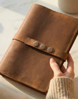 Brown leather notebook with button closure held by a hand on a light surface.