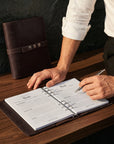 Person writing in a recipe book with a brown leather cover on a wooden table.