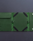 A dark green leather travel sleeve or tablet sleeve, shown open to reveal its interior compartments, elastic straps, and snap-button closure, set against a dark gray background.