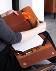 A person seated on a chair holds an open brown leather notebook cover or organizer on their lap. The cover contains lined paper, a pen loop with a pen, a card slot holding an orange card, and a small pocket.