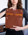 A woman holds a brown leather tablet sleeve or document holder with metal snap buttons, shown against an indoor background.