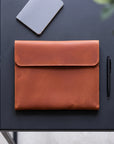 A tan leather envelope-style sleeve or pouch laid flat on a dark tabletop, with a gray notebook, black pen, and green leaves partially visible around it.