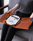 A person seated on a gray chair holds an open tan leather tablet organizer across their lap. The organizer contains a white tablet, two black pens, and a blue card, with black elastic straps securing the contents.