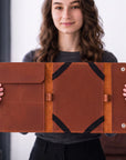 A woman holds open a large brown leather folio or tablet sleeve with black trim and snap-button closure, shown against a softly blurred indoor background.