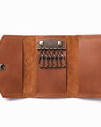 A brown leather key holder wallet shown open on a white background, featuring six metal key rings attached to a brass-tone clasp and a snap button closure on the left.