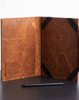 A brown leather folio or notebook cover shown open on a dark tabletop against a white background, with black corner straps and a small pen placed in front.