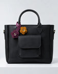A black leather handbag with dual top handles, a front flap pocket, and two decorative flower charms in purple and mustard yellow, displayed on a white pedestal against a light gray background.