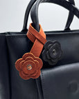 A close-up of a black leather handbag featuring two decorative flower-shaped leather charms hanging from the handle. One charm is a warm brown with a silver-tone center, and the other is black with a matching center.