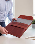 A person in a light blue dress shirt holds an open reddish-brown leather laptop sleeve with a closed silver laptop inside, set in a bright office environment with a desk, notebook, and plant in the background.