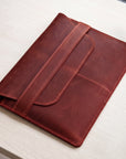 A reddish-brown leather folio or tablet sleeve with a strap closure, photographed on a light wood tabletop.