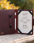 A burgundy leather tablet case shown open on a stone surface outdoors, with a white tablet inserted and black elastic bands holding it in place. The case has card slots on the left, a pocket tab, and a gold 'FB' monogram near the bottom left.