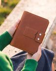 A person in a green sweater holds a brown leather journal or notebook with a snap-button strap closure, photographed outdoors on a sunny day.