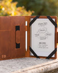 A brown leather tablet case stands open on a stone surface outdoors, with a white tablet inserted and black elastic bands holding it open. The case has card slots, a vertical pocket, and an embossed 'FB' logo, with a whiteboard-style screen area showing notes.