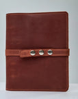 A leather recipe book with a green cover, secured with a snap closure, displayed on a table with a wrapped gift and a cake in the background.