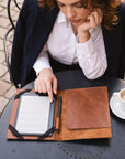 A woman in a navy blazer and white shirt sits at an outdoor table, looking down at an open brown leather folio case containing an e-reader, a pen, and a closed matching leather cover. A cup of coffee is on the table beside her.