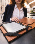 A woman seated at an outdoor cafe table uses a brown leather folio case with a black strap to hold an e-reader and a pen. The background shows a blurred street scene with buildings and cafe seating.