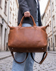 Person holding a brown leather duffel bag on a cobblestone street.