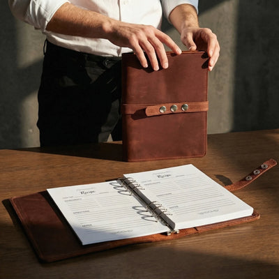 Brown leather recipe book with a person holding it open on a wooden surface.