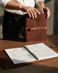 Brown leather recipe book with a person holding it open on a wooden surface.