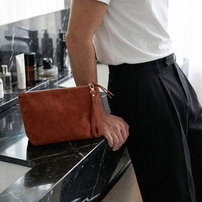 Person holding a brown clutch bag on a black marble surface with skincare products in the background.