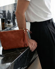 Person holding a brown clutch bag on a black marble surface with skincare products in the background.