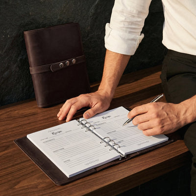 Person writing in a recipe book with a brown leather cover on a wooden table.