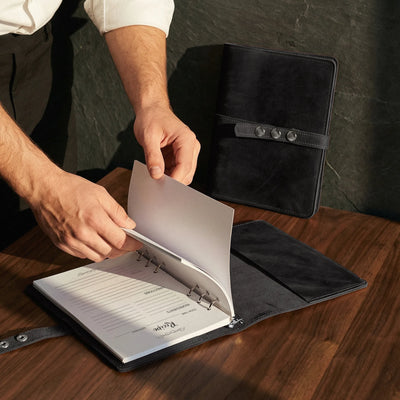 Person opening a black leather-bound notebook on a wooden surface