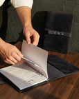 Person opening a black leather-bound notebook on a wooden surface