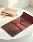 Brown leather wallet on a white surface with beige envelopes and a vase in the background.
