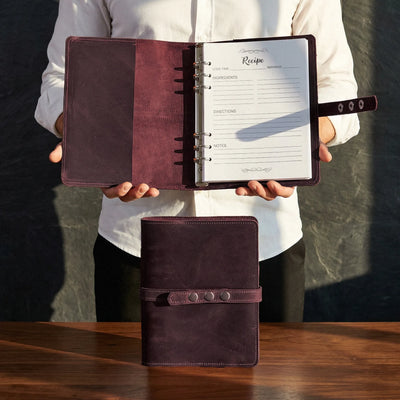 Person holding a brown leather planner with open pages on a wooden surface