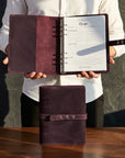 Person holding a brown leather planner with open pages on a wooden surface