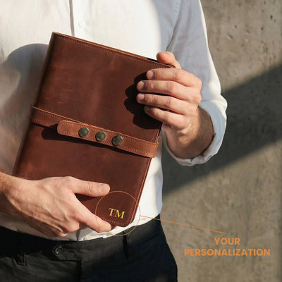 Person holding a brown leather notebook with 'TM' branding, against a neutral background.