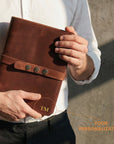 Person holding a brown leather notebook with 'TM' branding, against a neutral background.