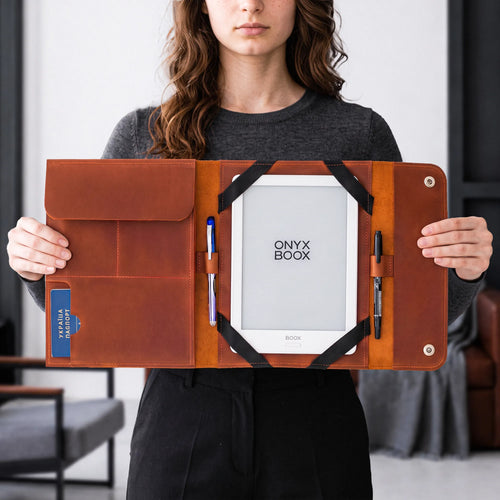 A woman holds open a brown leather folio-style tablet sleeve with a white Onyx Book e-reader inside, plus pens in pockets, against an indoor room background.