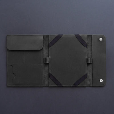 A dark gray leather tablet sleeve or folio case, shown open to display its interior compartments, elastic straps, and snap-button closure on a dark gray background.