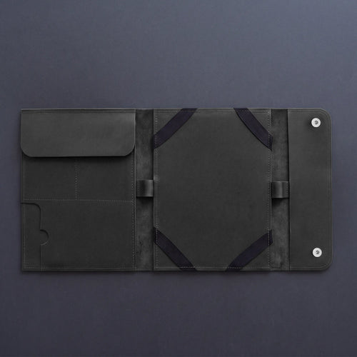 A dark gray leather tablet sleeve or folio case, shown open to display its interior compartments, elastic straps, and snap-button closure on a dark gray background.