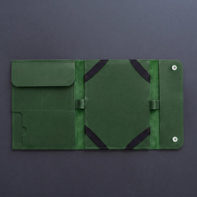 A dark green leather travel sleeve or tablet sleeve, shown open to reveal its interior compartments, elastic straps, and snap-button closure, set against a dark gray background.