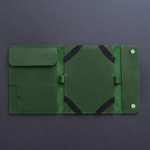 A dark green leather travel sleeve or tablet sleeve, shown open to reveal its interior compartments, elastic straps, and snap-button closure, set against a dark gray background.
