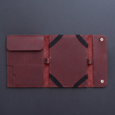 A dark reddish-brown leather travel organizer sleeve, shown open to reveal multiple pockets, elastic bands, and snap-button closure on a dark gray background.