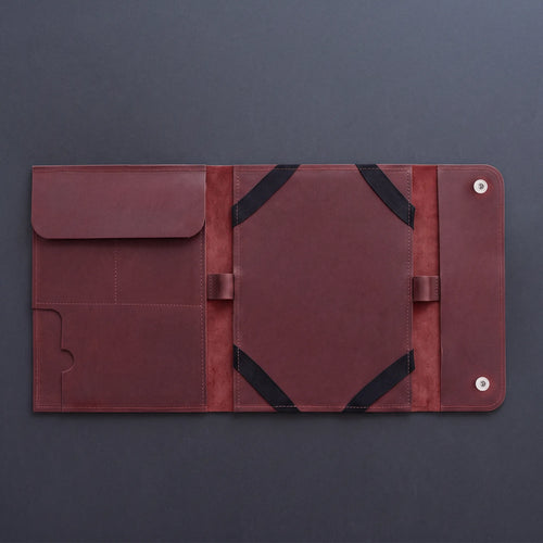 A dark reddish-brown leather travel organizer sleeve, shown open to reveal multiple pockets, elastic bands, and snap-button closure on a dark gray background.