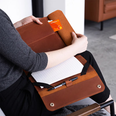 A person seated on a chair holds an open brown leather notebook cover or organizer on their lap. The cover contains lined paper, a pen loop with a pen, a card slot holding an orange card, and a small pocket.