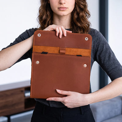 A woman holds a brown leather tablet sleeve or document holder with metal snap buttons, shown against an indoor background.