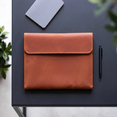 A tan leather envelope-style sleeve or pouch laid flat on a dark tabletop, with a gray notebook, black pen, and green leaves partially visible around it.