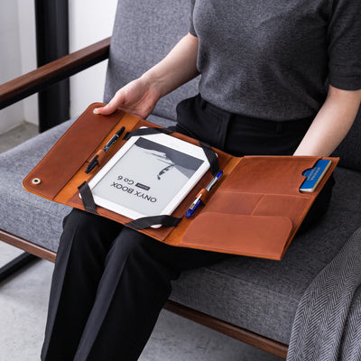 A person seated on a gray chair holds an open tan leather tablet organizer across their lap. The organizer contains a white tablet, two black pens, and a blue card, with black elastic straps securing the contents.