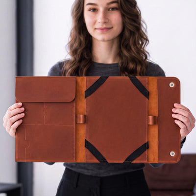 A woman holds open a large brown leather folio or tablet sleeve with black trim and snap-button closure, shown against a softly blurred indoor background.