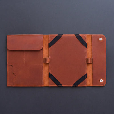 A brown leather organizer sleeve or folio, shown open to reveal multiple pockets, elastic bands, and snap-button closure, set against a dark gray background.