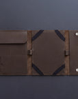 A dark brown leather travel sleeve or document holder, shown open to display its interior compartments, elastic straps, and snap-button closure on a dark gray background.