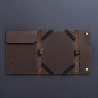 A dark brown leather travel sleeve or document holder, shown open to display its interior compartments, elastic straps, and snap-button closure on a dark gray background.