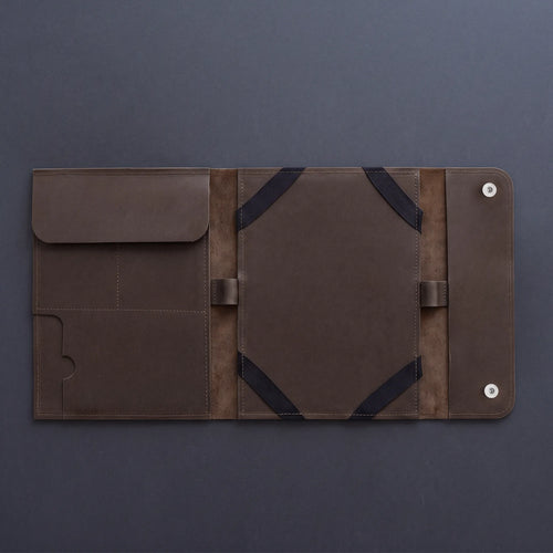 A dark brown leather travel sleeve or document holder, shown open to display its interior compartments, elastic straps, and snap-button closure on a dark gray background.