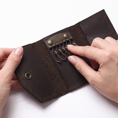 A close-up of a dark brown leather key holder or key ring roll being opened by two hands, showing brass-toned metal key rings and snap buttons on a white background.